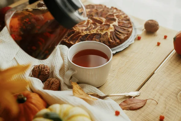 Cozy autumn. Pouring herbal tea in stylish cup on background of wooden table with freshly baked warm apple pie with cinnamon, autumn leaves, pumpkins and nuts. Hygge home