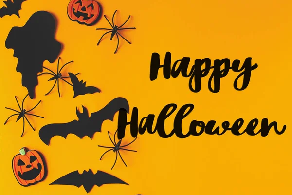 Happy Halloween text on bats, spiders, ghosts and pumpkins flat lay on yellow background. Happy Halloween greeting card, handwritten sign. Trick or treat!