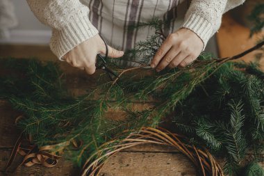 Kırsal Noel çelengi yapıyorum. Köknar dallarını kırsal ahşap bir masanın arka planında kurdeleyle kesen kadın eller. Huysuz Noel imajı, tatil hazırlıkları