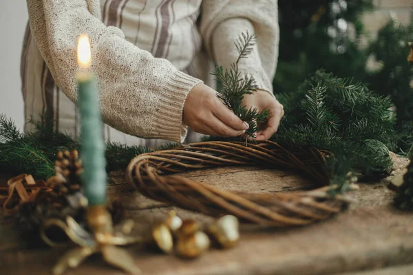 クリスマスの素朴な花輪を作る 女性の手は リボン 黄金の鐘 ろうそくと素朴な木製のテーブルの上にモミの枝を保持し 花輪を作る ムーディーの休日のイメージ 冬休みワークショップ — ストック写真