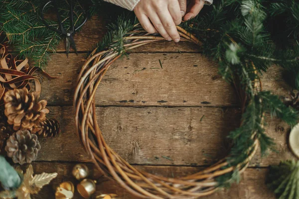 Making rustic Christmas wreath. Fir branches on round wicker wreath, pine cones, ribbon, golden bells and candles on rustic wooden table. Moody Christmas still life, holiday preparations