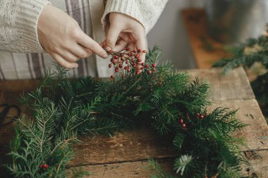 Noel çelengi yapıyorum. Kırsal masada kırmızı böğürtlen tutan ve Noel çelenginde köknar dalları süsleyen kadın elleri. Huysuz tatil imajı. Tatil atölyesi, kış dekorasyonu.