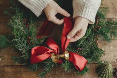 Noel çelengi yapıyorum. Kadın elleri Noel çelengini kırmızı kurdeleyle süslüyor ve çam kozalakları ve mumlarla ahşap masa üzerinde altın çanlar çalıyor. Huysuz tatil imajı. Tatil atölyesi
