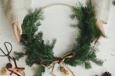 Modern Noel çelengi düzdü. Elleri şık bir Noel çelengi, kadife kurdele ve beyaz ahşap masada altın çanlar tutuyor. Kış tatili dekorasyonu, karamsar bir görüntü. Mutlu Noeller.!