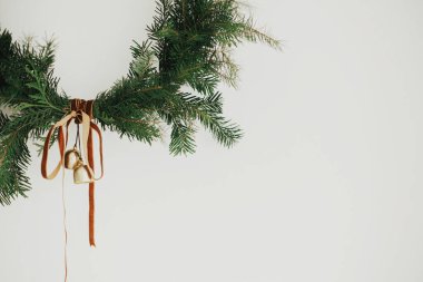 Beyaz arka planda kadife kurdeleli ve altın zilli modern Noel çelengi. Mutlu Noeller ve mutlu tatiller! Kış tatili dekorasyonu, karamsar bir görüntü. Boho çelengi