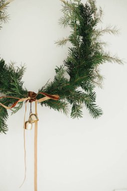 Kadife kurdeleli modern Noel çelengi ve beyaz duvarda asılı altın çanlar yakın plan. Mutlu Noeller ve mutlu tatiller! Kış tatili dekorasyonu, karamsar bir görüntü. Boho çelengi