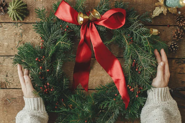 Woman holding stylish christmas wreath with red ribbon, golden bells and berries on rustic wooden table flat lay. Making Christmas wreath, moody image. Winter holiday decor. Merry Christmas!