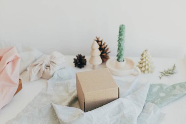 Furoshiki hediye paketi. Hediye kutusu, keten tekstil, tahta ağaç oyuncağı, beyaz ahşap masada mum. Sıfır Noel konsepti. Çevre dostu yeniden kullanılabilir Noel hediyesini şık kumaşla sarma