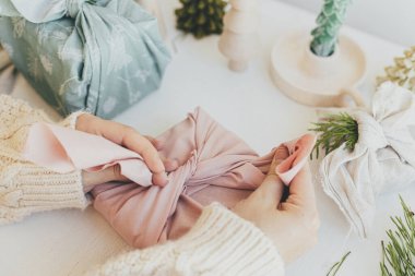 Furoshiki hediye paketi. Beyaz ahşap masa üzerinde pembe keten kumaşlarla hediye paketleri, çevre dostu ahşap oyuncaklar, köknar dalı, mum. Sıfır Noel konsepti. Kış hazırlıkları