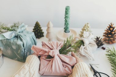 Furoshiki hediye paketi. Elleri pembe kumaşa sarılı, köknar dallı hediye paketi beyaz ahşap masa üzerinde, çevre dostu dekor ve mum ile. Sıfır Noel konsepti. Mutlu Noeller.!