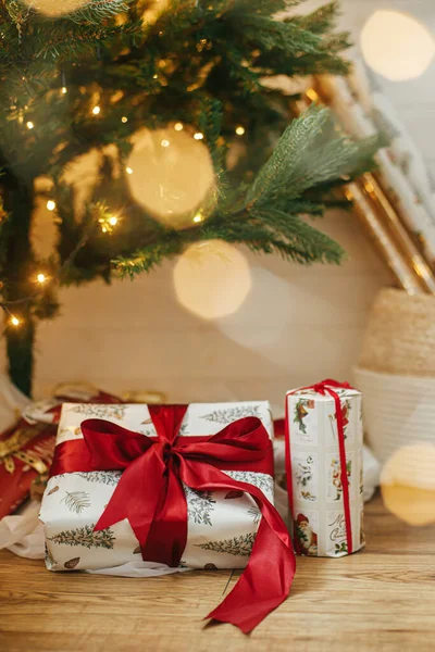 金色のライトボケとクリスマスツリーの下でスタイリッシュなクリスマスプレゼント 部屋の照明と装飾された木の下に赤いリボンで包まれたクリスマスプレゼント 大気圏イブ ホリデーバナー — ストック写真