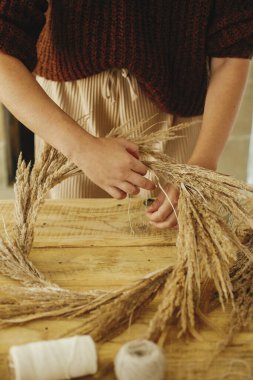 Elleri çember üzerine kurumuş çim düzenleme tahta masa üzerinde makas, iplik. Kırsal masada şık bir sonbahar çelengi yapmak. Çiftlik evinde sonbahar dekorasyonu ve düzenleme.
