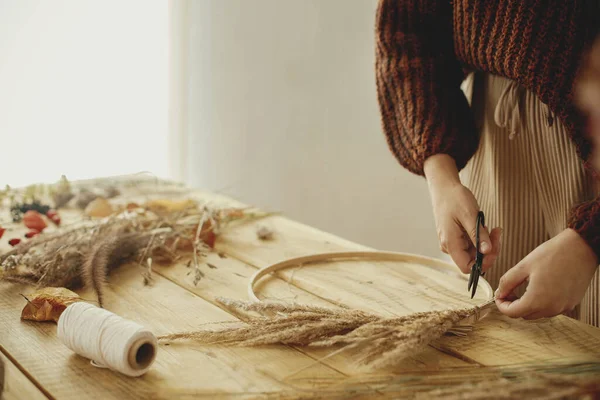 Hands arranging dried grass on hoop on wooden table with scissors, thread. Making stylish autumn wreath on rustic table. Fall decor and arrangement in farmhouse.