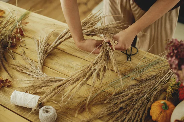 Hands arranging dried grass on hoop on wooden table with scissors, thread. Making stylish autumn wreath on rustic table. Fall decor and arrangement in farmhouse.