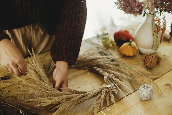 Woman hands arranging dried grass in wreath on wooden table with scissors, thread, pumpkin. Making stylish autumn wreath on rustic table. Fall decor and arrangement in farmhouse.