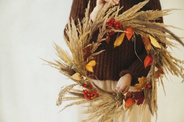 Kurumuş çimen, böğürtlen, otlar ve yapraklarla süslü bir sonbahar çelengi. Beyaz duvar arkasında kırsal çelenk tutan şık bir kadın. Çiftlik evinde sonbahar dekorasyonu ve düzenleme