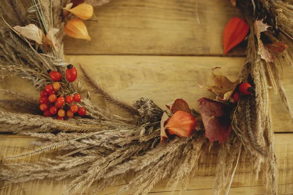 Rustic autumn wreath with dried grass, berries, herbs on wooden table. Fall decor and arrangement in farmhouse. Thanksgiving. Details of stylish autumn wreath on rustic table close up