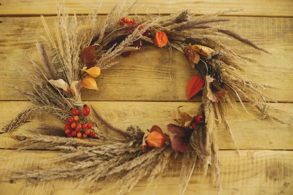 Rustic autumn wreath with dried grass, berries, herbs on wooden table. Fall decor and arrangement in farmhouse. Thanksgiving. Details of stylish autumn wreath on rustic table close up