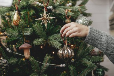Atmosferik festival odasında Noel ağacını şık takılarla süsleyen eller. Mutlu Noeller! Kış tatili hazırlığı. Samimi süveterli bir kadın ağaca antika bir oyuncak koyuyor.