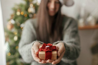 Işıklı yılbaşı ağacına karşı elinde şık bir Noel hediyesi. Mutlu Noeller ve mutlu tatiller! Sıcak kazaklı kadın hediyesini sarıp atmosferik festival odasına sarmaladı.