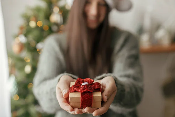 Stylish christmas gift in hands against christmas tree with lights. Merry Christmas and Happy Holidays! Woman in cozy sweater holding wrapped present close up in atmospheric festive room