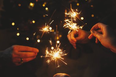 Mutlu yıllar! Karanlık odada Noel ışıklarına karşı havai fişek tutan eller. Atmosferik tatil. Arkaplanda yanan kıvılcımlarla kutlayan arkadaşlar şık ışıklandırılmış ağacın arkasında
