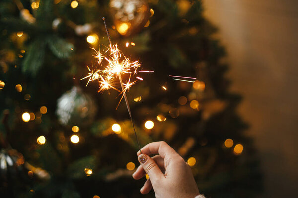 С Новым Годом! Горящий блеск в женской руке на фоне рождественских ёлочных огней в темной комнате. Атмосферное празднование. Ручной фейерверк против стильного украшенного дерева с подсветкой