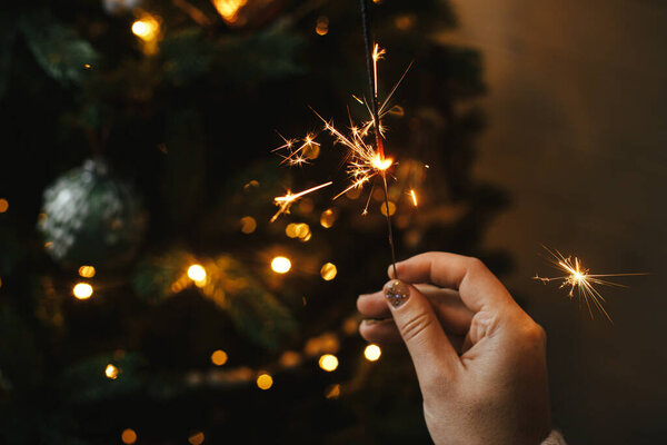 Hand holding firework against christmas tree lights in dark room. Happy New Year! Merry Christmas! Burning sparkler in female hand  on background of golden illumination bokeh. Atmospheric time