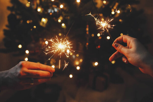 Руки держат фейерверк на рождественских ёлочках в темной комнате. С Новым Годом! Пара празднующих с горящими искрами в руках на фоне стильного декорированного дерева с подсветкой.