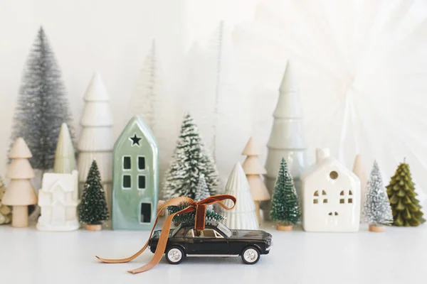 Holidays are coming! Stylish little car carrying christmas tree on background of Christmas miniature snowy village. Merry Christmas! Festive winter scene on white table, holiday banner