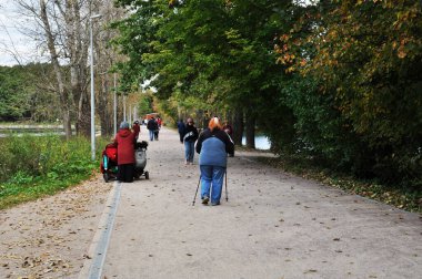 Şehir parkının panoraması. İnsanlar park yolu boyunca yürür. Bilinmeyen bir kadın İskandinav yürüyüşü yapıyor..