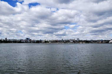 Büyük gölün panoramik manzarası ve konut binaları ile kıyı şeridi. Gökyüzünde beyaz bulutlar var..