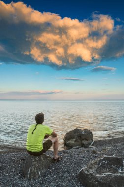 Deniz kenarındaki bir taşın üzerinde oturan ve gökyüzündeki günbatımı bulutlarına bakan bir adam..