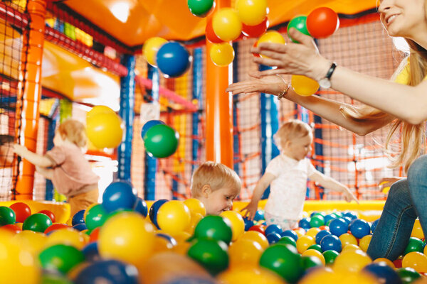 Happy kids playing in amusement park balls pool with a young teacher. Kids cheering and playing with plastic colourful balls. Children throwing balls high. Playing with balls: activities for children