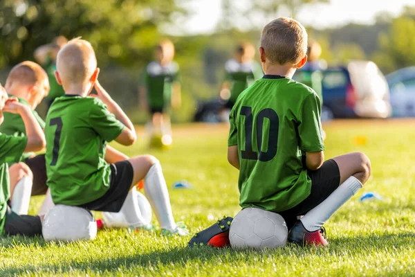 若い男の子のグループ草のピッチ上のスポーツサッカーゲームキッキング 少年たちはサッカーボールに座ってゲームをプレイするのを待っている スポーツチームの友人 シドラインの男子生徒 グリーンスポーツシャツの子供たち — ストック写真