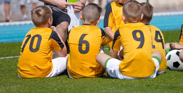 フットボールの少年たちのコーチ戦術のアドバイスを聞いてのグループ サッカーのピッチでトレーナーと座っている学校の子供たち 監督はユースチームに試合の計画を示す ユースチームでのサッカー指導 — ストック写真