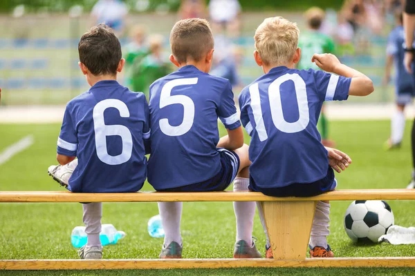 青年队的足球队员坐在木制长椅上 学校运动队的孩子们身穿蓝色球衣的男孩观看比赛 — 图库照片