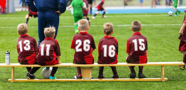学校足球队女巫教练坐在木制长椅上观看比赛 穿着红衫的孩子们准备踢足球 — 图库照片