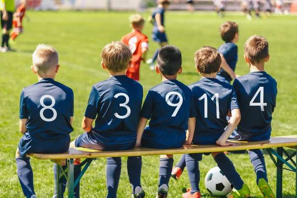 青いジャージーのシャツを着たサッカー選手たちが木のベンチに腰を下ろしている 学校のスポーツチームの子供たちの多人種グループ 青いジャージーのシャツの男の子はトーナメントゲームを見て — ストック写真
