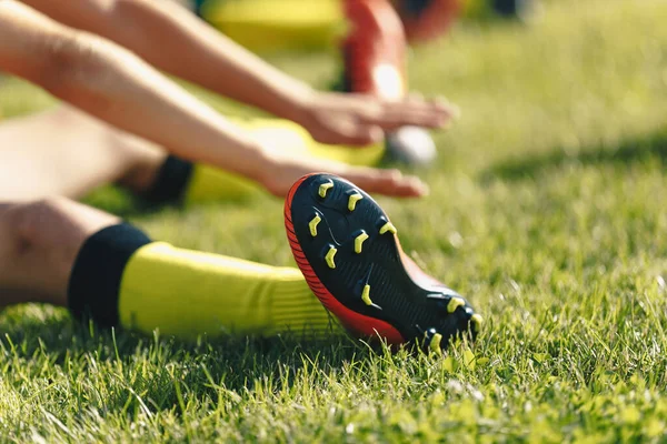 Football Player Stretches Sitting on Grass Pitch. Stretching Session After Workout For Footballers. Player in Soccer Cleats and Socks