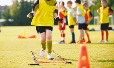 School Boy Running Slalom Track Between Training Poles and Jumping Over Ladders. Teenage Football Players Running in Two Rows on Training Camp. Soccer Training Equipment