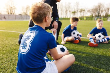 Happy children sitting on football field with soccer balls and listening to school coach. Young coach teaching kids on football field. 