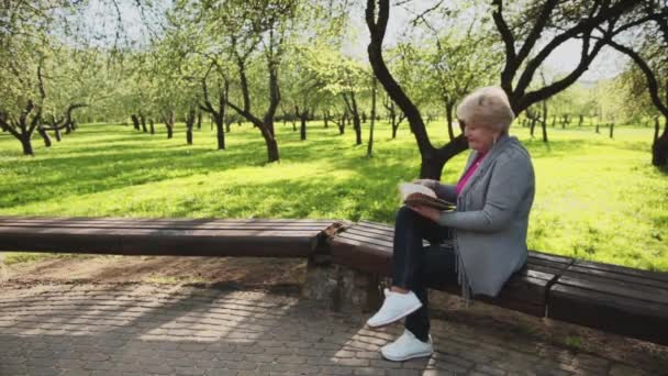 En kvinna sitter på en parkbänk och läser en bok.. — Stockvideo