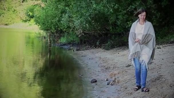 Задумчивая женщина, завернутая в одеяло прогулки на берегу реки — стоковое видео