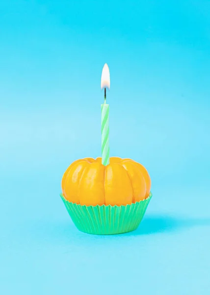 Lighted birthday candle on ripe orange pumpkin in green cupcake cup against sky blue background.