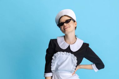 Oda hizmetçisi üniforması ve gözlüklü genç bir kadın mavi arka planda izole edilmiş.