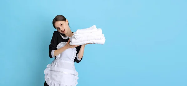 Ung attraktiv kammarjungfru i uniform med fastnat färska handdukar talar på telefon isolerad över blå bakgrund. Flygblad — Stockfoto