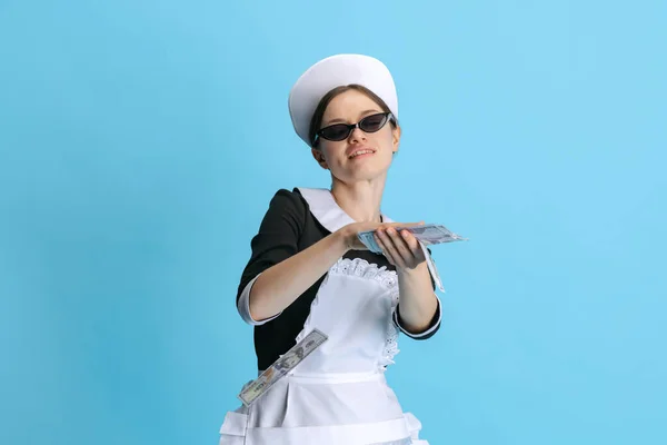 Mujer joven y mandona en uniforme de camarera y gafas de sol elegantes aisladas sobre fondo azul —  Fotos de Stock