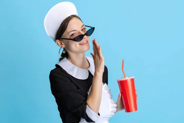 Menina bonita vestindo uniforme de camareira e bebendo refrigerante isolado sobre fundo azul — Fotografia de Stock