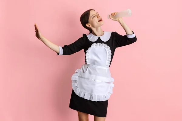 Portret młodej, pięknej pokojówki hotelowej w czarnym mundurze śpiewającej w sprayu do czyszczenia butelki na różowym tle. — Zdjęcie stockowe